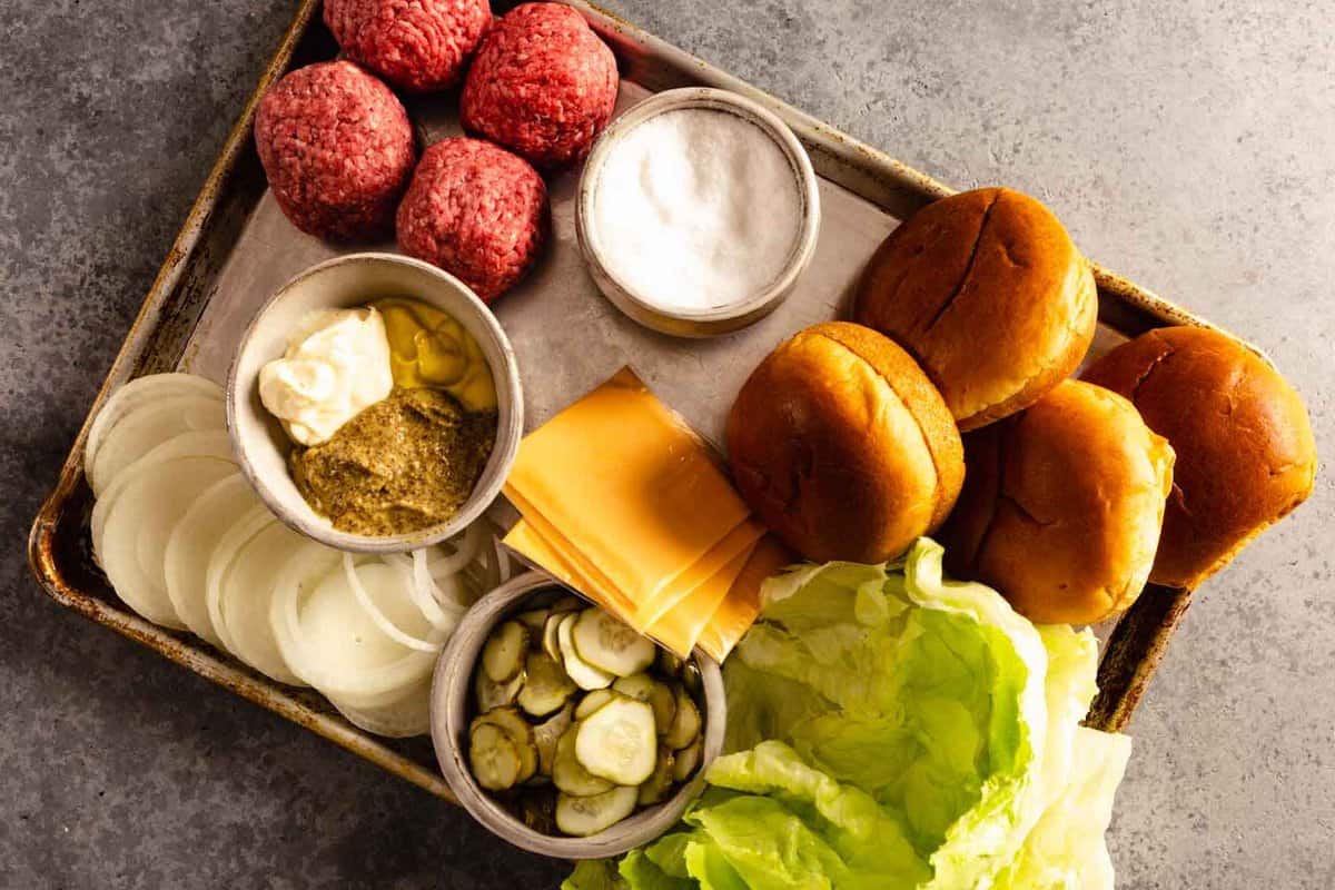 balls of ground beef, burger buns, lettuce, pickles, cheese, mustard and mayo, and sliced onions arranged on a baking sheet