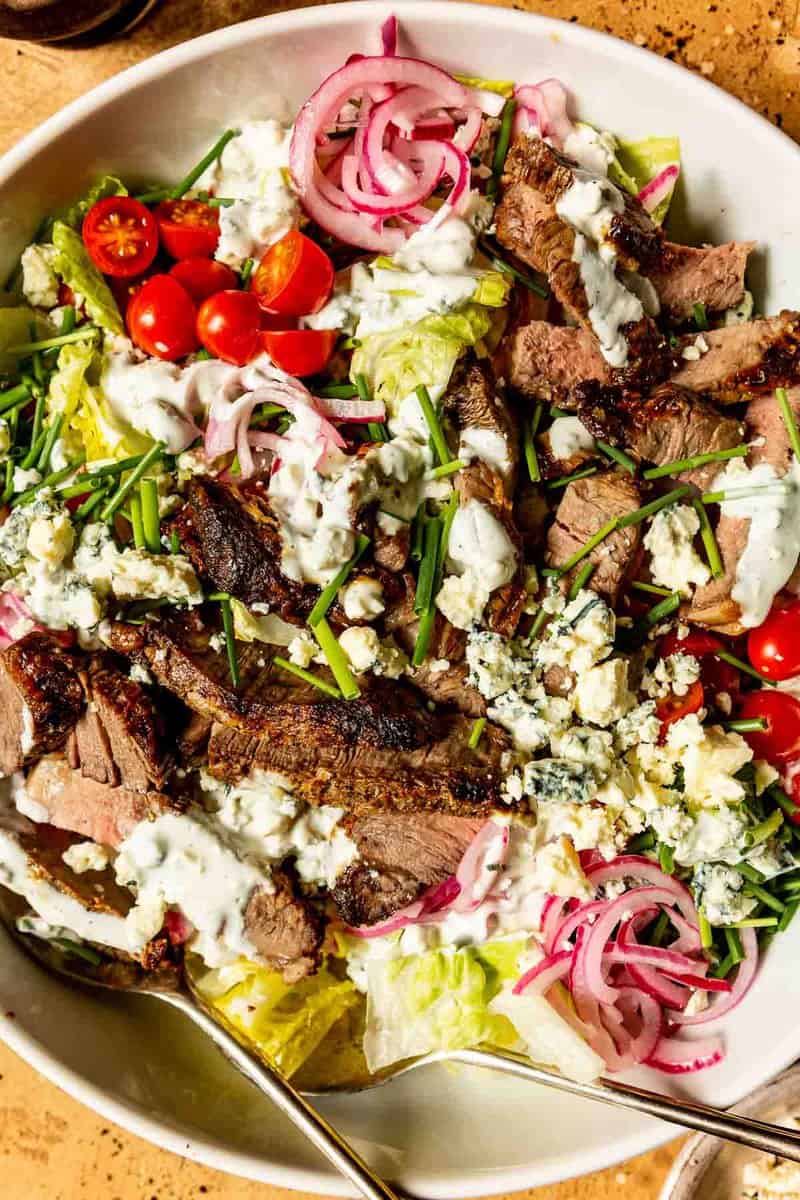 blue cheese steak salad in a large white bowl with romaine lettuce, cherry tomatoes, pickled onions, chives, sliced steak and a creamy dressing drizzled over it all