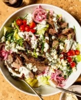 blue cheese steak salad in a large white bowl with romaine lettuce, cherry tomatoes, pickled onions, chives, sliced steak and a creamy dressing drizzled over it all