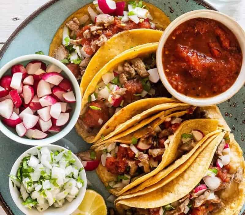 4 steak tacos arranges on a teal of plate with salsa and toppings set around them