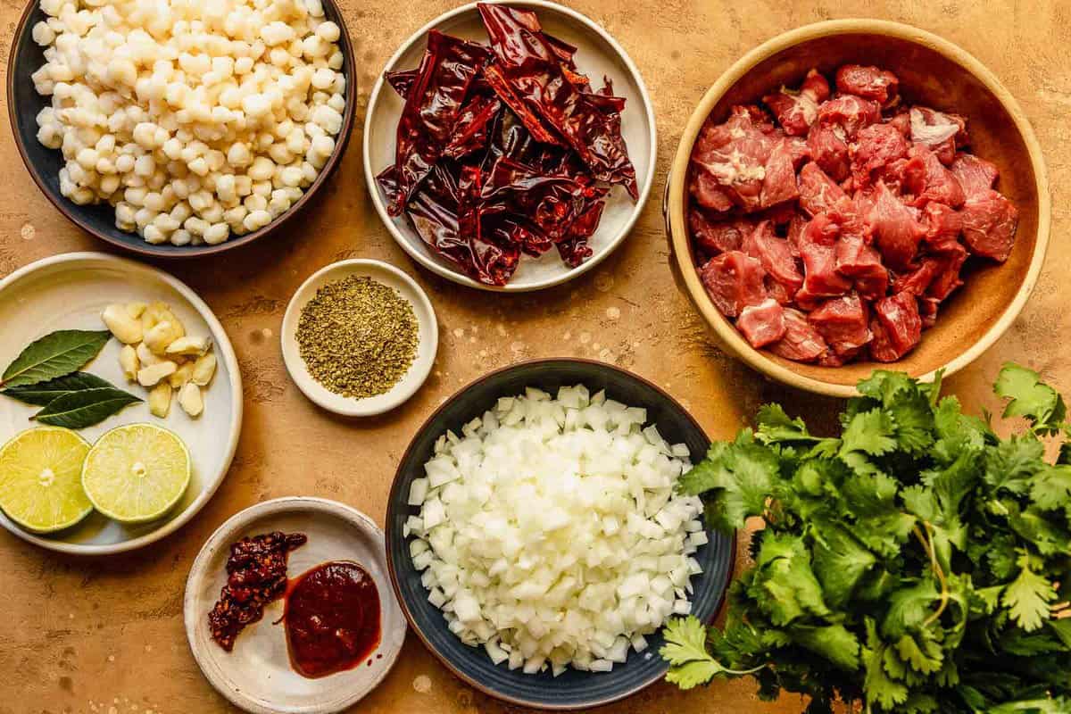 raw cubed pork, dried chiles, diced onions, hominy, cilantro, chipotle in adobo, dried oregano, and garlic measured out and set on a counter