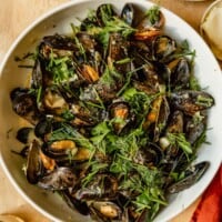 mussels in a large white bowl with chunks of bread sitting in the broth, glasses of white wine set around it
