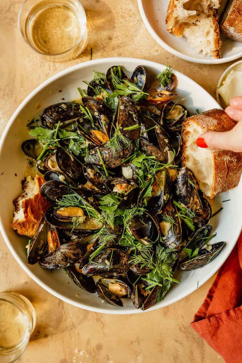 mussels in a large white bowl with chunks of bread sitting in the broth, glasses of white wine set around it