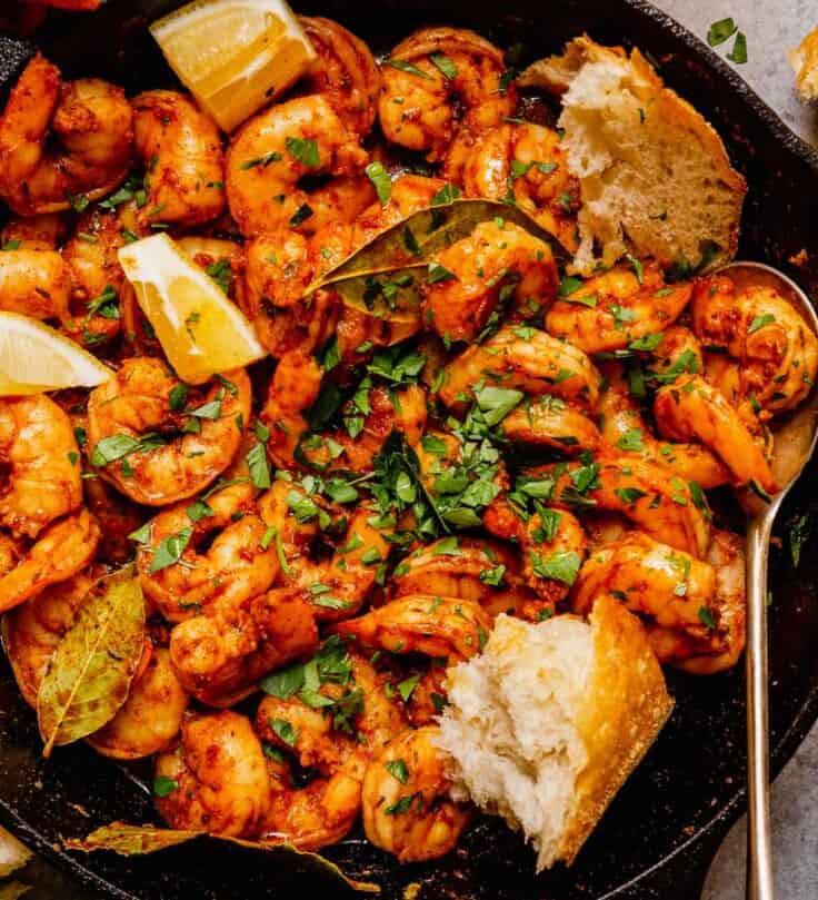 spiced shrimp in a cast-iron skillet with hunks of bread, lemon wedges and chopped parsley