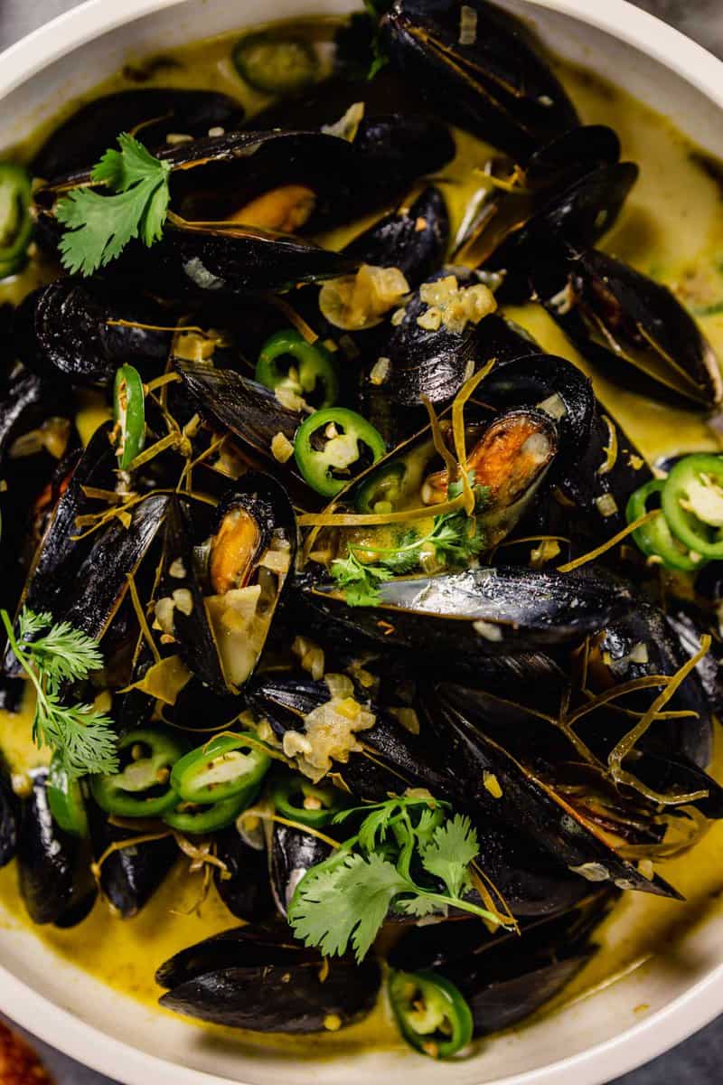 steamed mussels in a large white bowl with a creamy yellow broth and green herbs