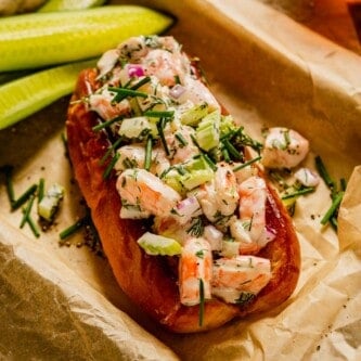 shrimp salad piled into a brioche bun set in a parchment-lined baking sheet with cucumber slices