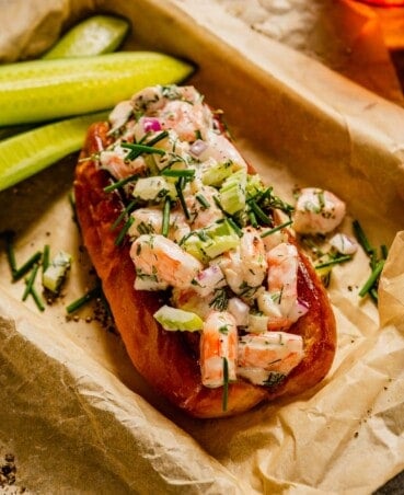 shrimp salad piled into a brioche bun set in a parchment-lined baking sheet with cucumber slices