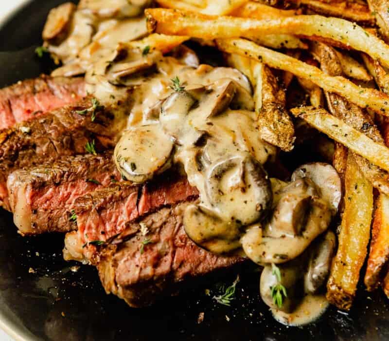 creamy mushroom sauce on top of sliced medium rare steak on a plate with fries