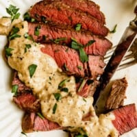 sliced steak on a white plate topped with a creamy sauce and parsley