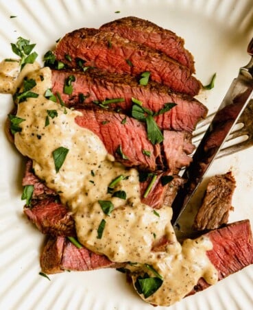 sliced steak on a white plate topped with a creamy sauce and parsley