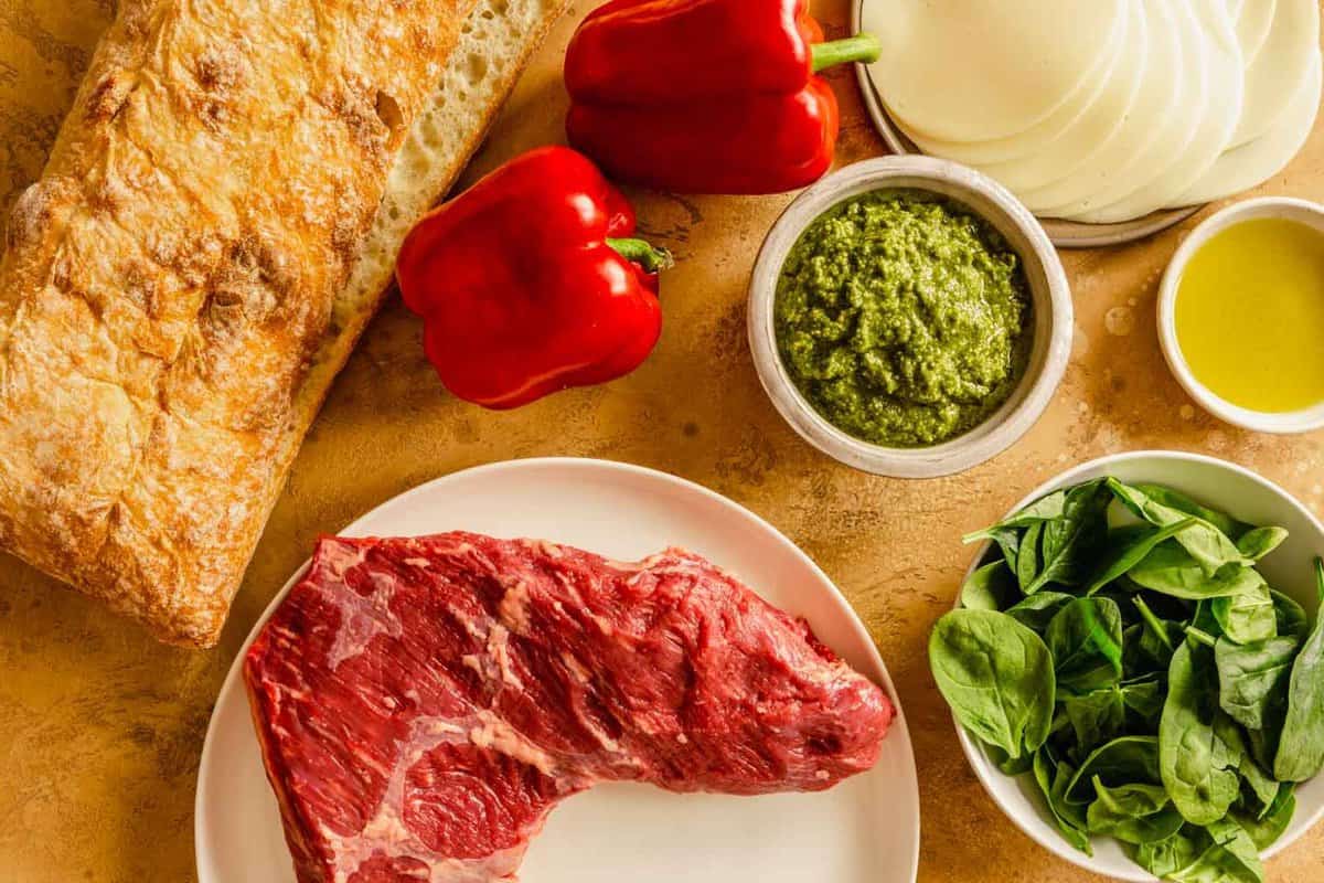 olive oil, spinach, cheese, pesto, red peppers, ciabatta, and steak arranged on a counter