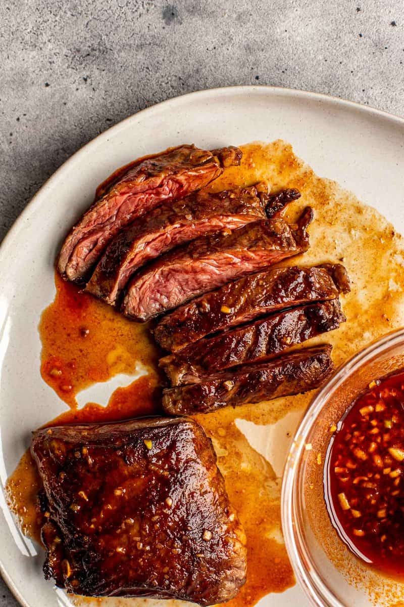 sliced steak on a plate with red sauce brushed over steak and in a bowl set next to steak