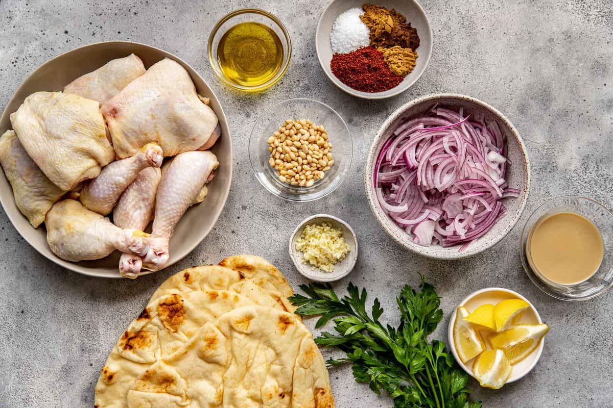 Raw bone-in, skin-on chicken thighs, sliced red onions, olive oil, a bowl with mixed spices, naan, parsley, pine nuts, minced garlic and lemon wedges set out on a counter.