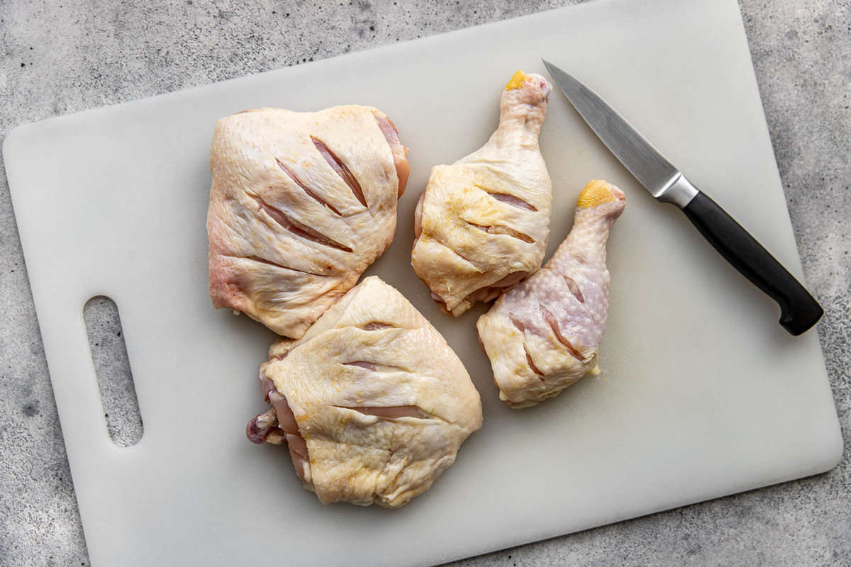 Raw bone-in, skin-on chicken thighs and drumsticks on a plastic cutting board with slits cut in them a knife set to the side.