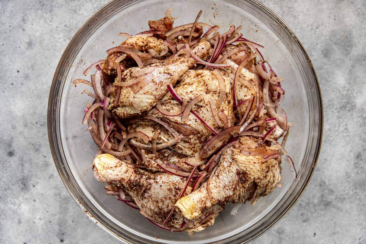Raw bone-in, skin-on chicken thighs and drumsticks coated in a spice paste in a bowl with sliced red onions.