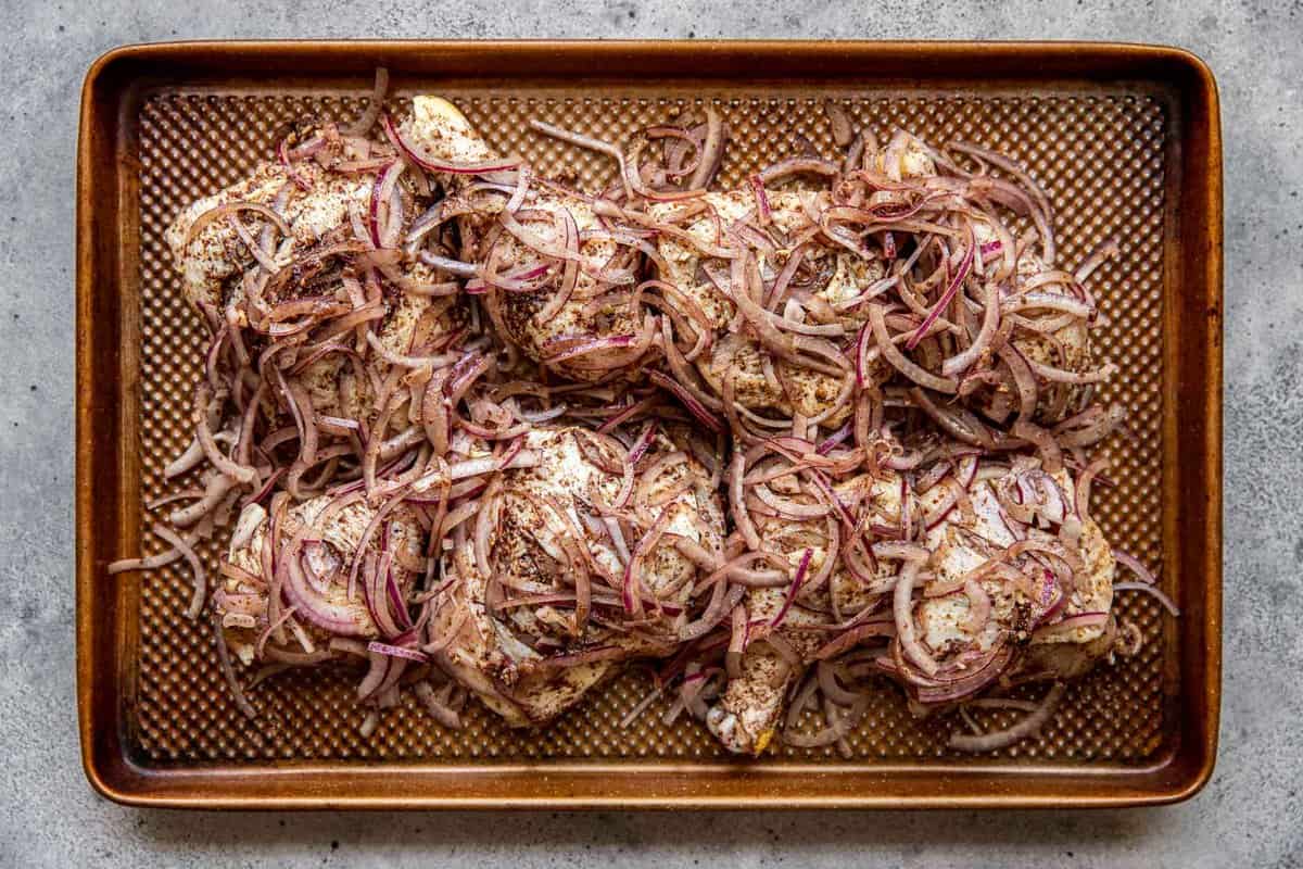 Raw bone-in, skin-on chicken thighs and drumsticks coated in a spice paste and topped with sliced red onions on a baking sheet.