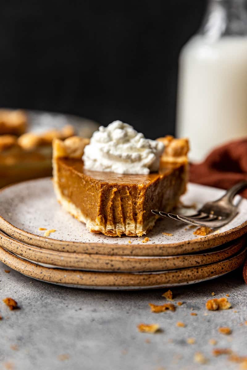 slice of pumpkin pie on a speckled plate with a fork and topped with whipped cream. Tip of piece removed with fork tine imprint on filling.