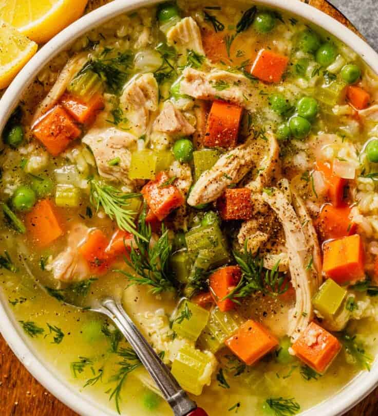 Brothy chicken and rice soup with diced vegetables, fresh dill and lemon in a shallow white bowl.