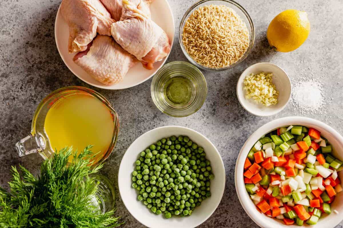 Ingredients for lemon chicken rice soup set out on a counter including chicken thighs, rice, chicken broth, vegetables, lemon, peas and dill. 