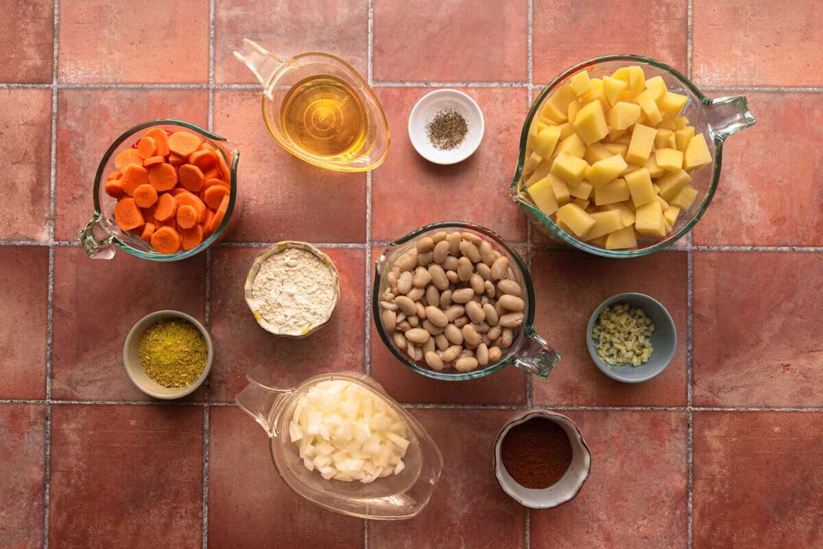 ingredients for serbian bean soup set out on a counter. Prepped ingredients include diced potatoes, carrots, and onions along with Vegeta, olive oil, paprika, white beans, salt and pepper.