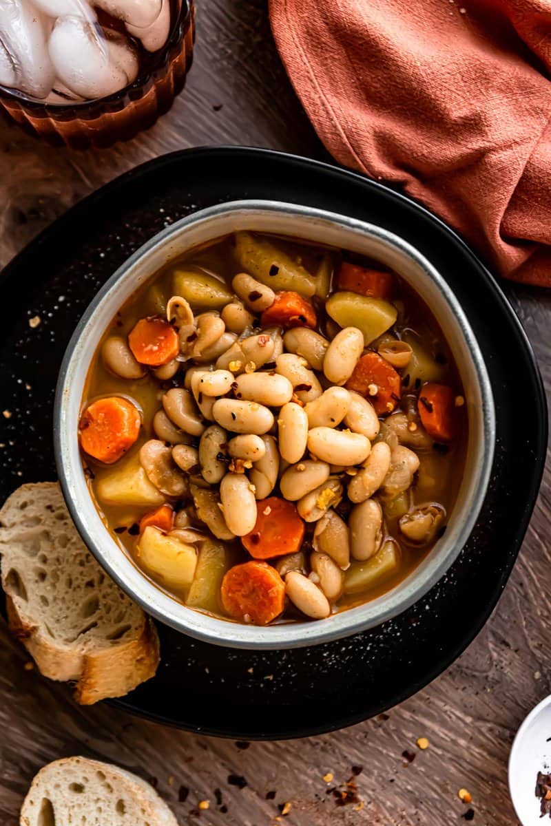 Serbian white bean soup in a gray bowl set on a black plate with a crusty piece of bread.