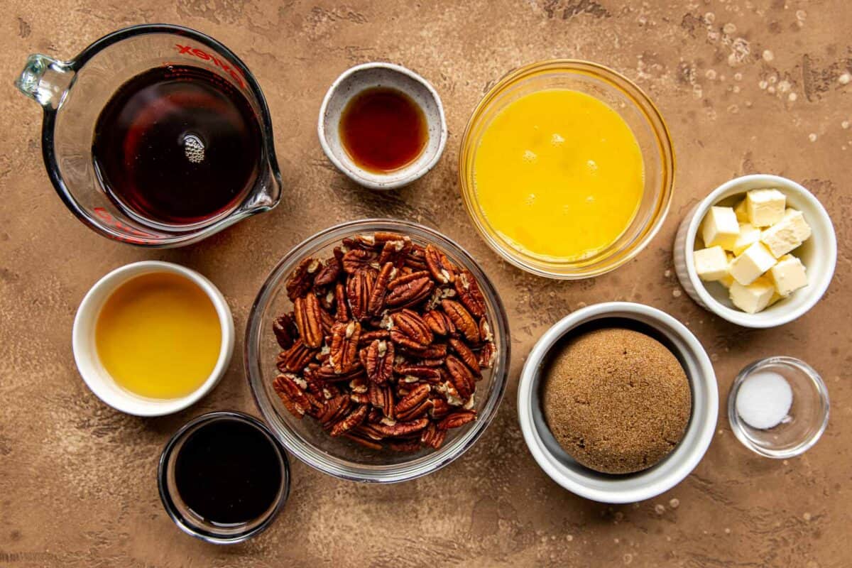 Bourbon pecan pie filling ingredients set out on a counter in clear glass bowls. Bowls filled with maple syrup, dark brown sugar, molasses, beaten eggs, pecan halves, butter, and salt.