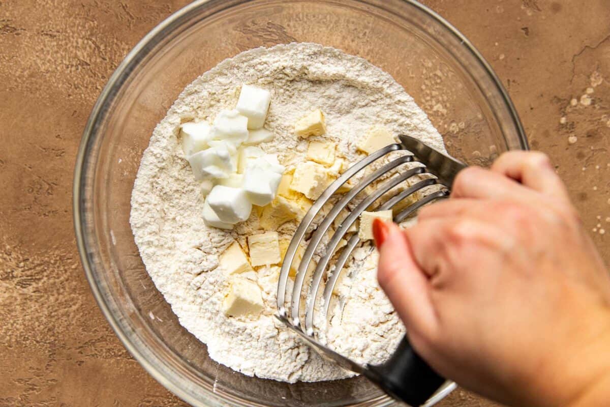 Someone's hand using a pastry blender to cut butter and shortening into a flour mixture in a glass bowl.