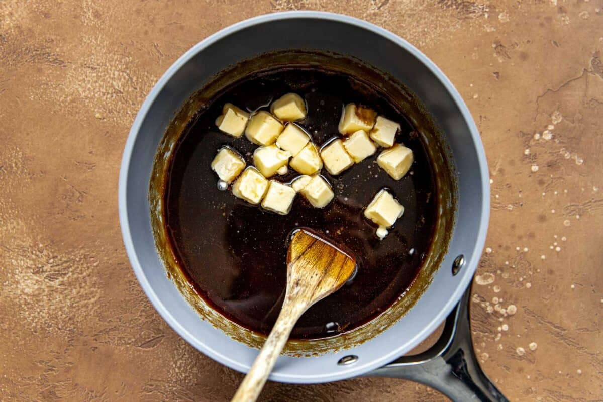 Cubes of butter in a saucepan of maple syrup, brown sugar, and molasses with a wooden spoon.