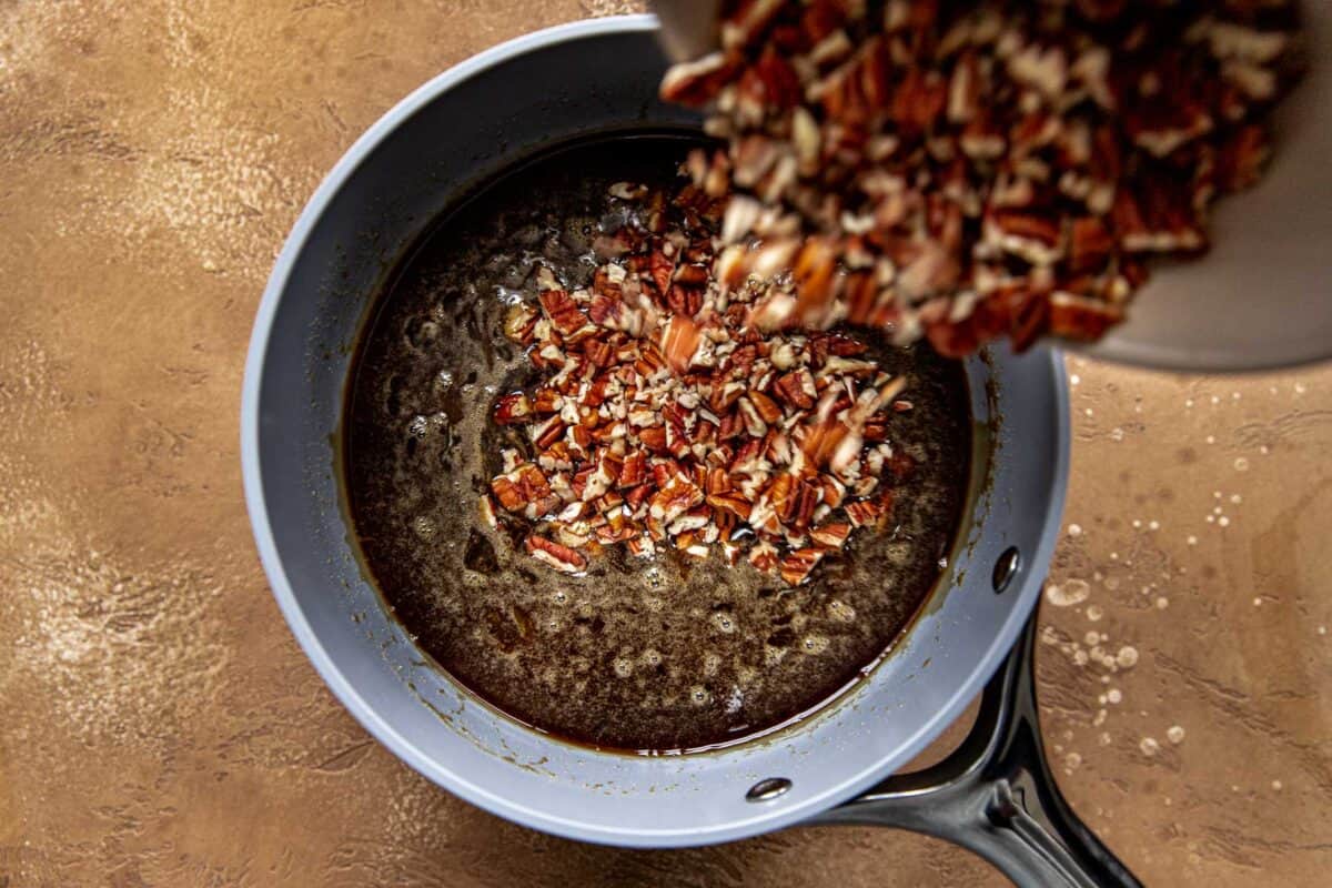 Chopped pecans being pouring into a maple syrup and sugar mixture in a saucepan.