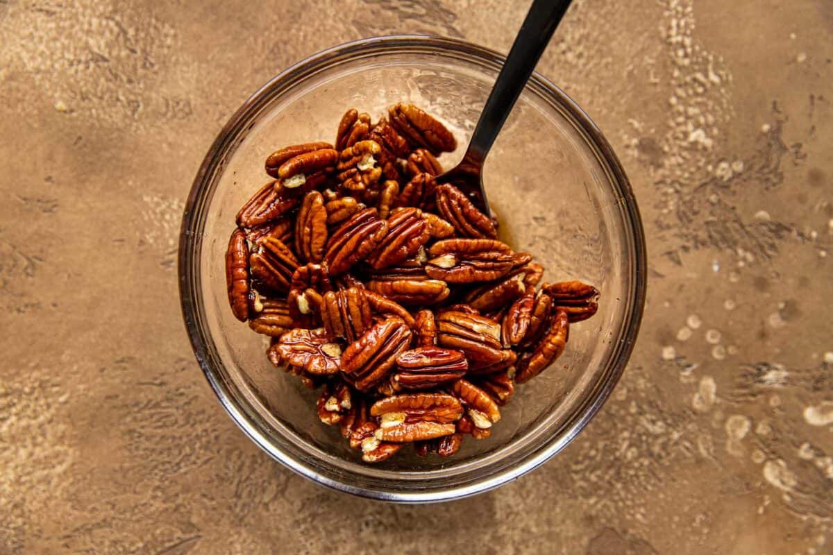 Pecan halves tossed with maple syrup and bourbon in a small glass bowl with a spoon.
