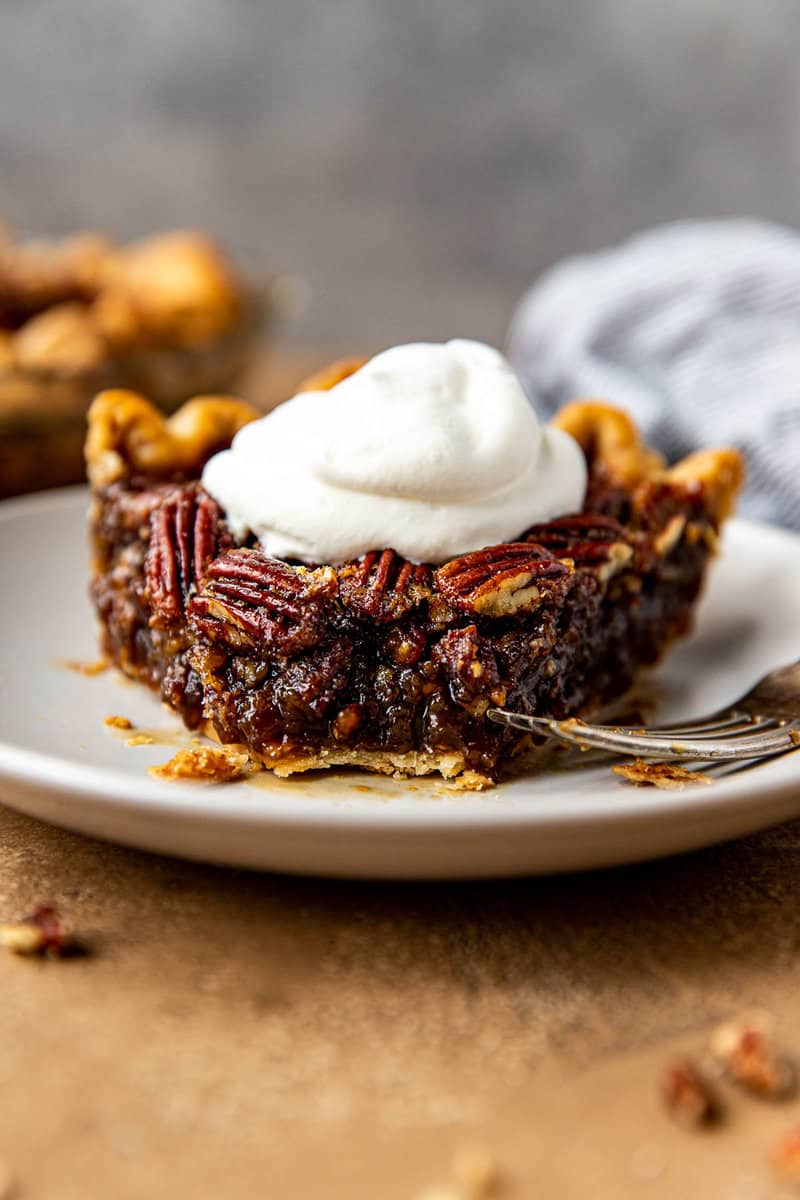 Slice of boozy pecan pie on a white plate with a fork; whipped cream on top.