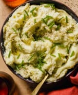 Boursin mashed potatoes in a shallow blue bowl with olive oil, chives and black pepper scattered over top. Dish set on a light orange table with orange glasses set around.