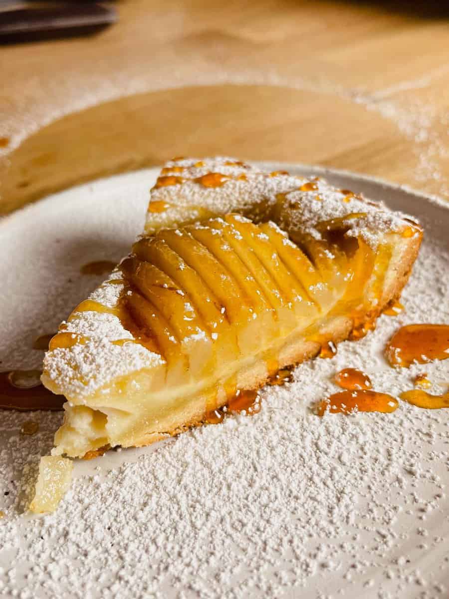 a slice of pear tart with almond filling and poached pears on a white  plate topped with caramel sauce and powdered sugar.
