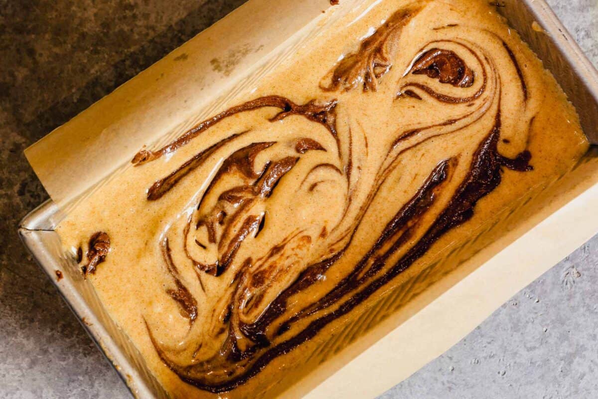 Hazelnut cake batter in a loaf pan with chocolate swirled into it.