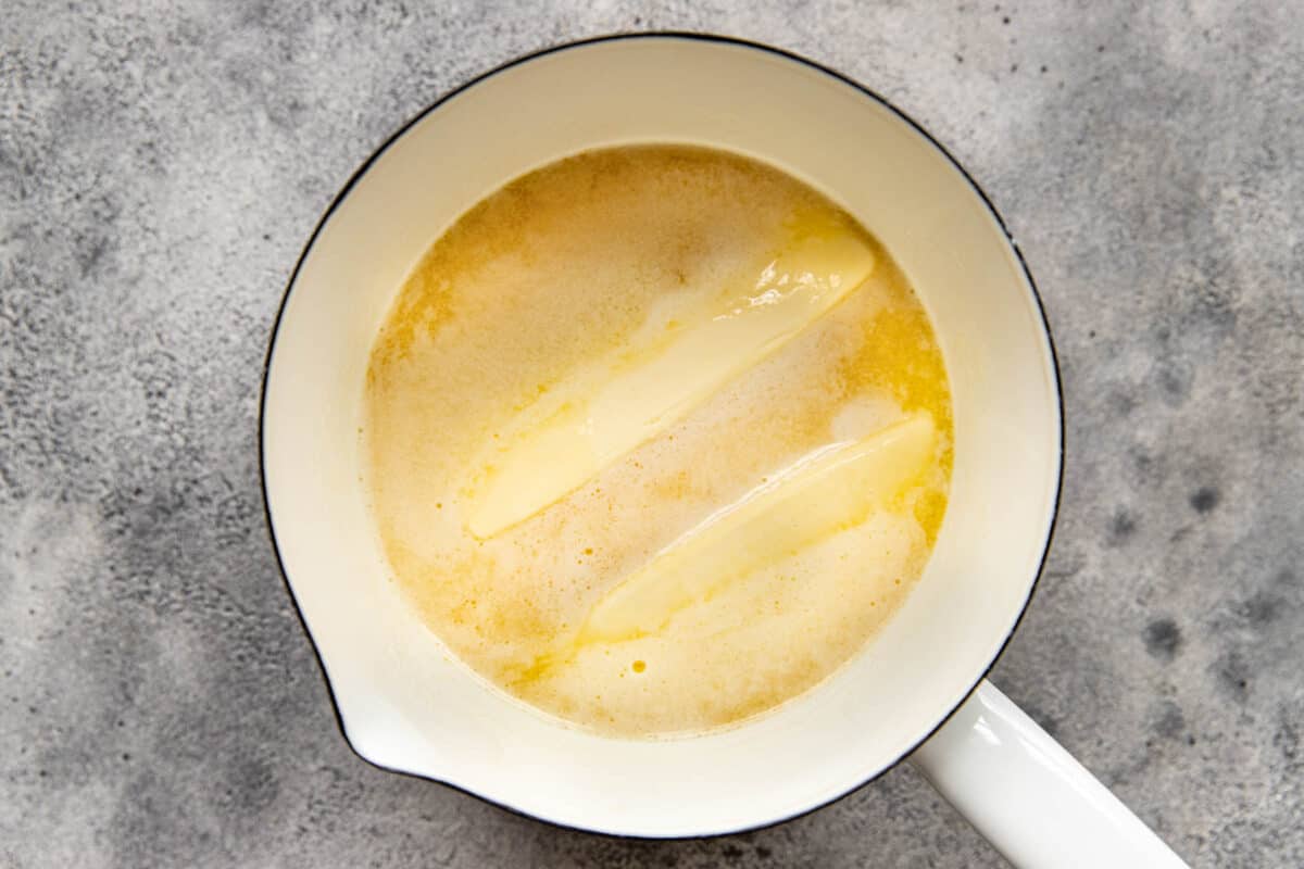 two sticks of butter melting in a white saucepan.