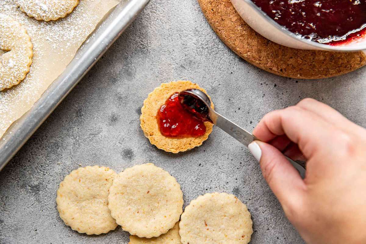 jelly being spooned onto linzer cookies with a half teaspoon measure.