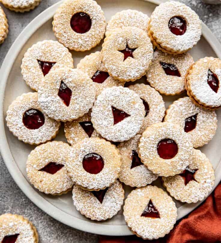 Linzer cookies filled with a red jam and stacked on a round platter with an orange napkin set to the side.