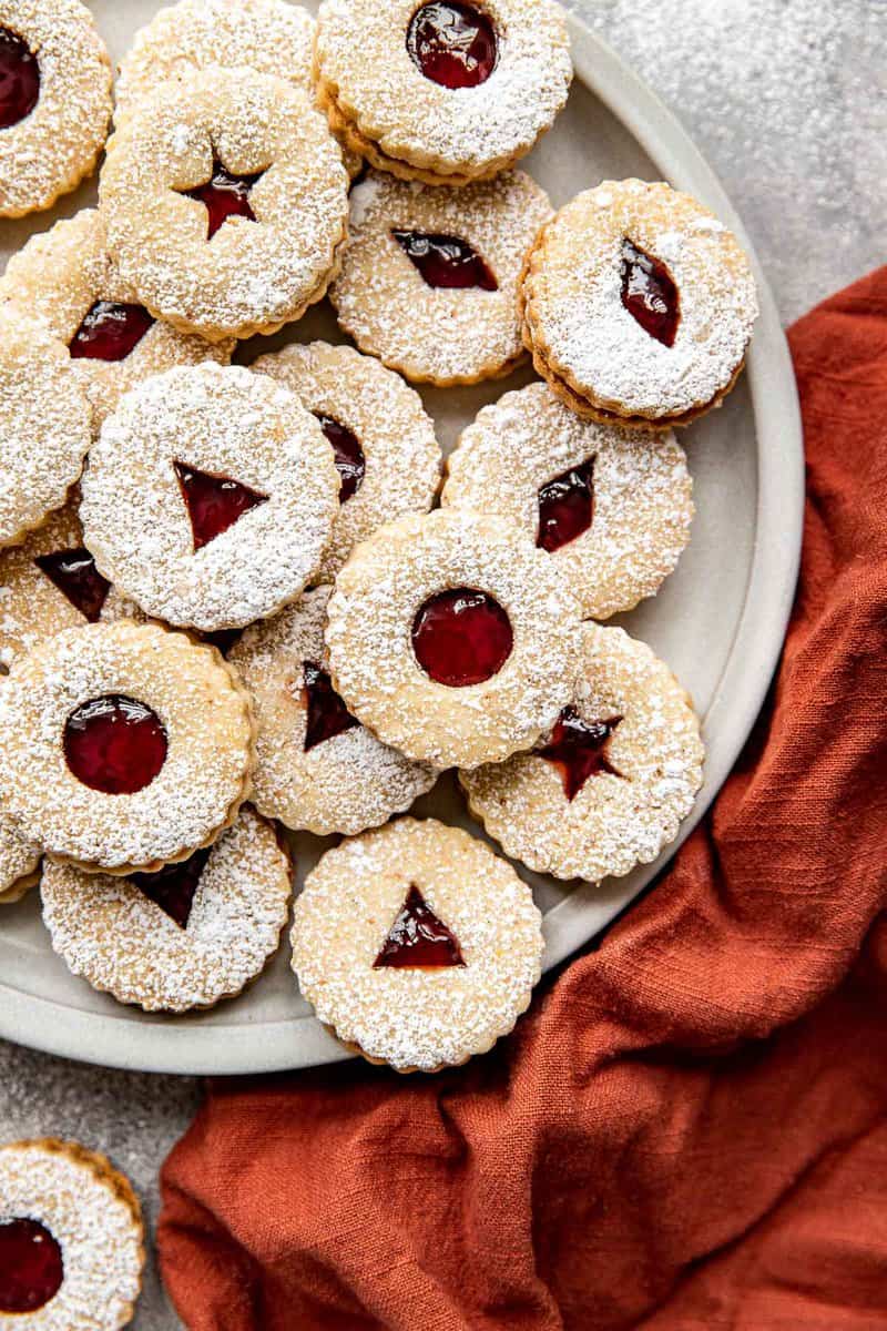 Linzer cookies filled with a red jam and stacked on a round platter with an orange napkin set to the side. 