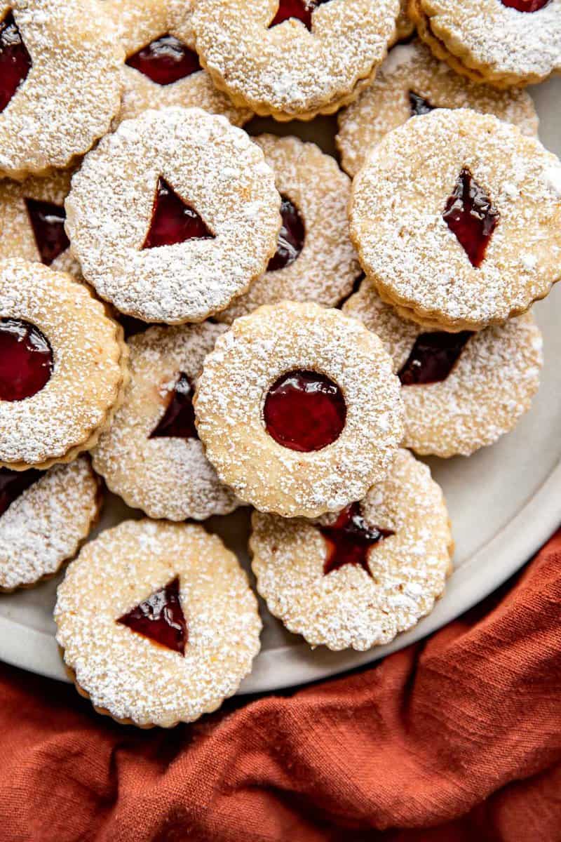Linzer cookies filled with a red jam and stacked on a round platter with an orange napkin set to the side.