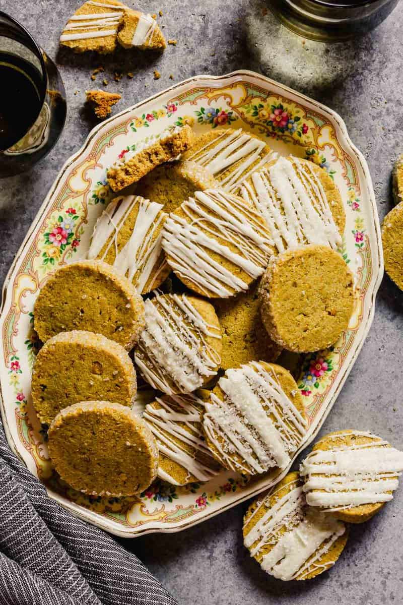 Pistachio shortbread cookies stacked on a floral platter. Some cookies drizzled with white chocolate, some with edges covered in sparkling sugar and some with nothing on them.