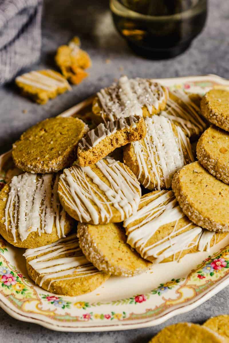 Pistachio shortbread cookies stacked on a floral platter. Some cookies drizzled with white chocolate, some with edges covered in sparkling sugar and some with nothing on them.
