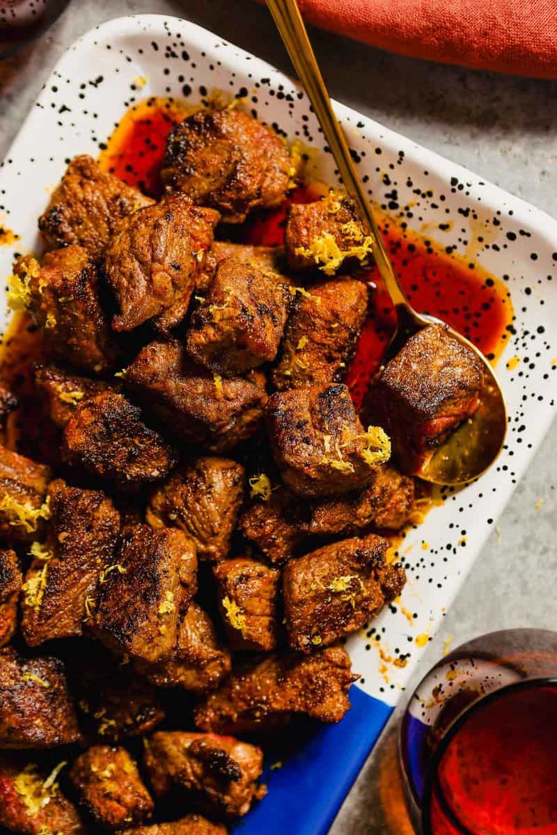 Steak bites on a speckled white, black and blue platter with a gold spoon.