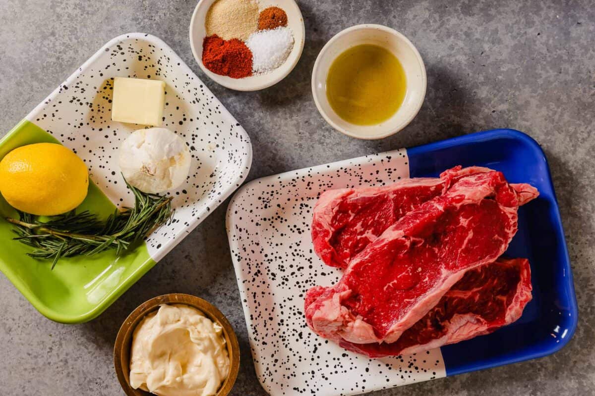 two large steaks, olive oil, spices, butter, rosemary and mayo set out on a counter.