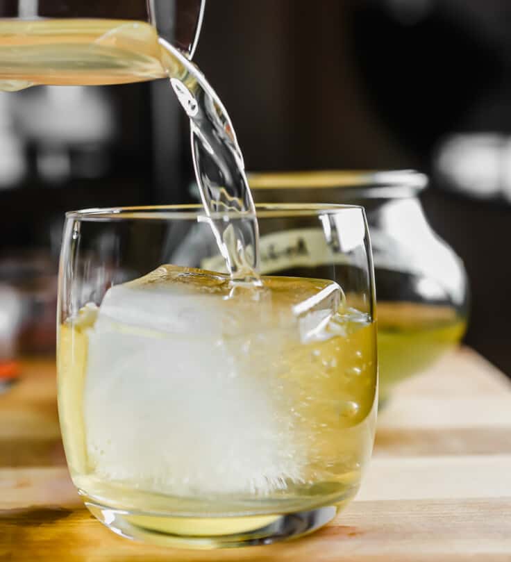 Clear yellow clarified milk punch cocktail being poured over a large ice cube in a rocks glass.