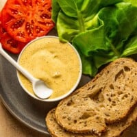 Dijon mustard in a small white bowl set on a plate with thick slices of bread, tomatoes and lettuce leaves.
