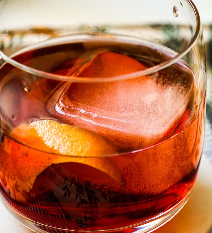 Red-hued non alcoholic spritz in a rocks glass with a large ice cube and orange peel. Cocktail set on a coaster on a patterned tray.