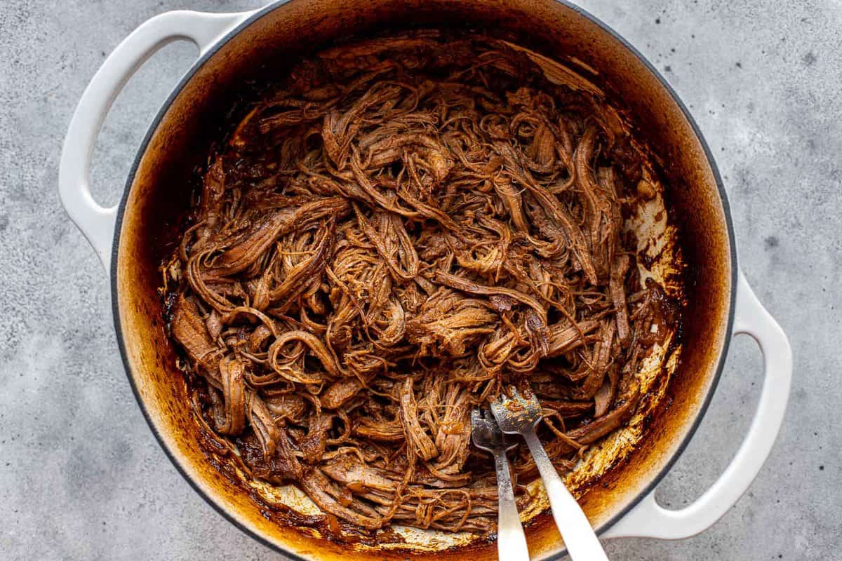 shredded saucy beef brisket in a large Dutch oven with two forks.