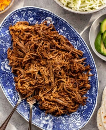 Mexican shredded beef piled on a large blue and white patterned oval platter with taco toppings set around.