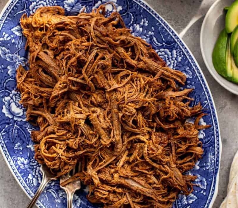 Mexican shredded beef piled on a large blue and white patterned oval platter with taco toppings set around.