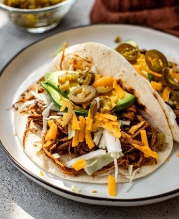 Shredded beef tacos on a plate topped with shredded cheese, avocados, pickled jalapenos, and shredded cabbage.